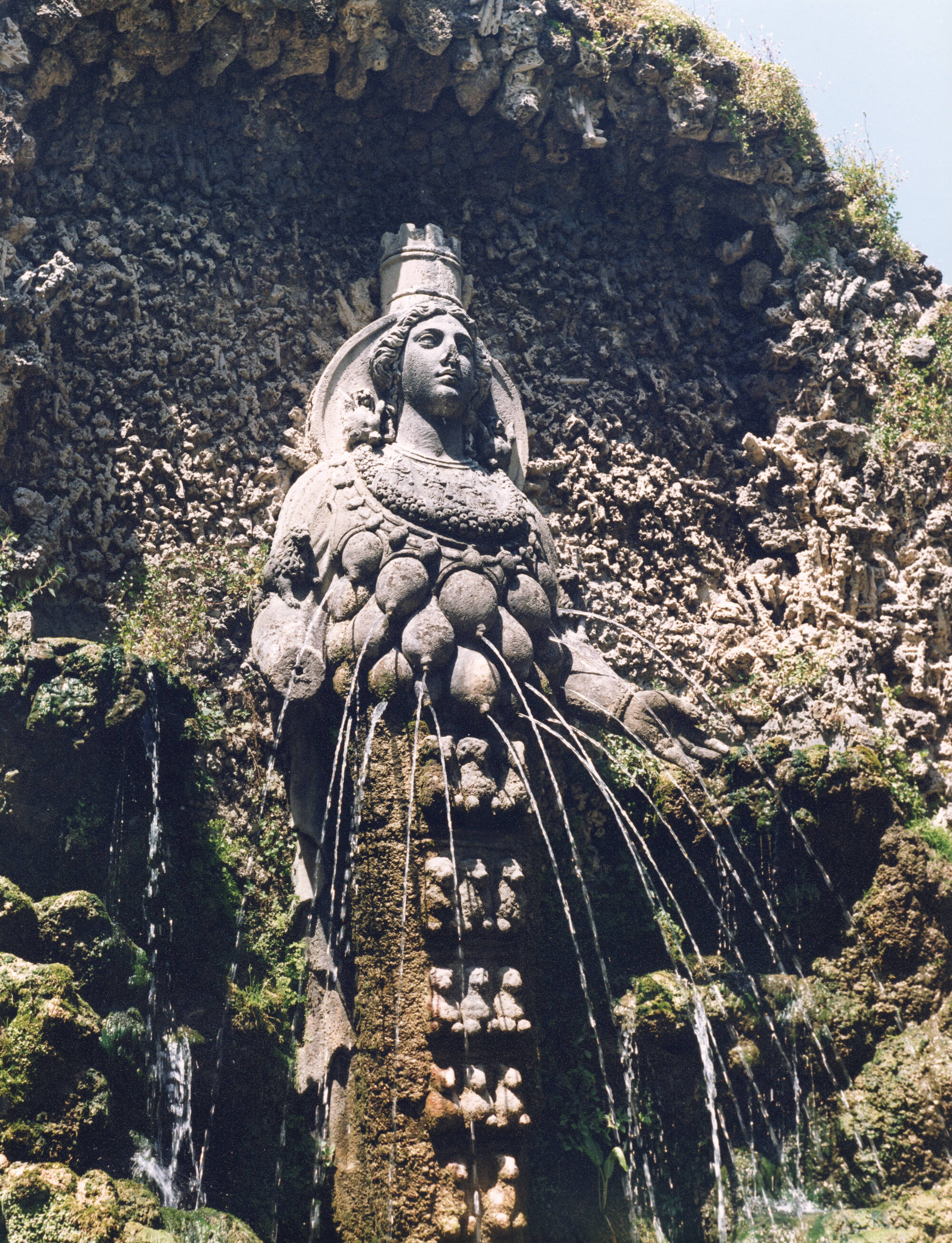 Photograph of a sculpture of a figure that is spraying out water out of multiple breasts on the torso. The sculpture is up against a stone wall that's growing greenery.